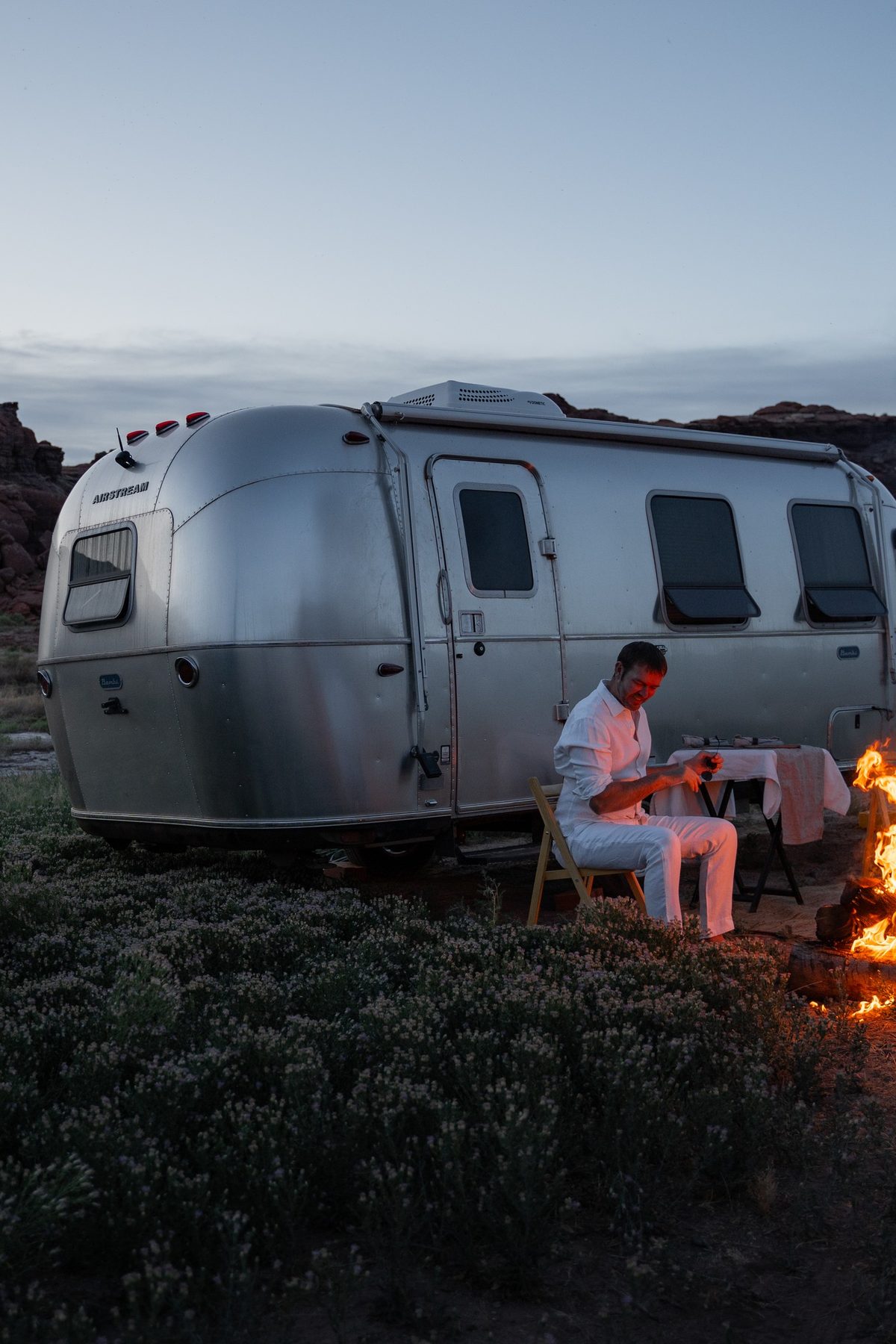 A guest by a campfire next to an Airstream trailer at dusk